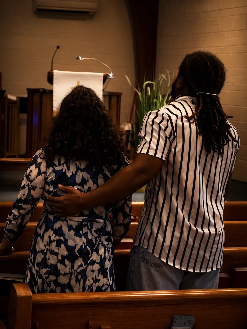 Two believers standing side by side in the pews during worship, one with an arm around the other